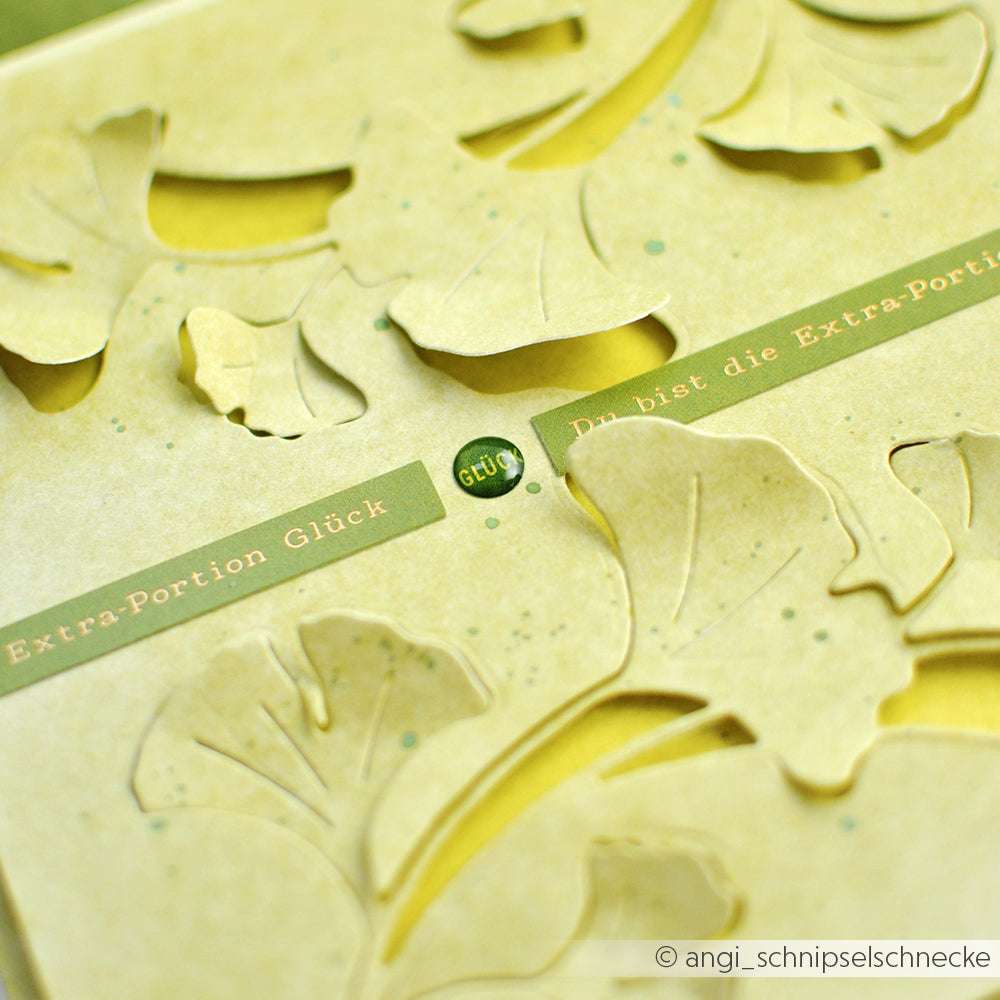 Nahaufnahme einer gelben Alexandra Renke-Karte mit ausgeschnittenen Blattformen und deutschen Textstreifen, darunter "ExtraPortion Glück" - ideal für kreative Kartengestaltung oder Verzieren mit Sticker-Streifen.