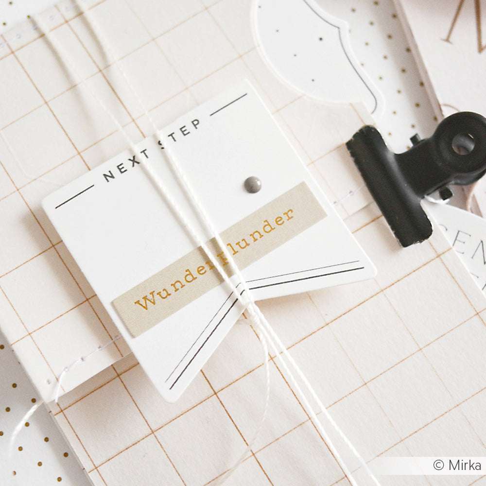 Ein Papieranhänger mit der Aufschrift "NEXT STEP WunderWunder" von Alexandra Renke, ideal für die Kartengestaltung, an einer Schnur auf gitterförmigem Hintergrund befestigt und mit einer schwarzen Klammer fixiert.