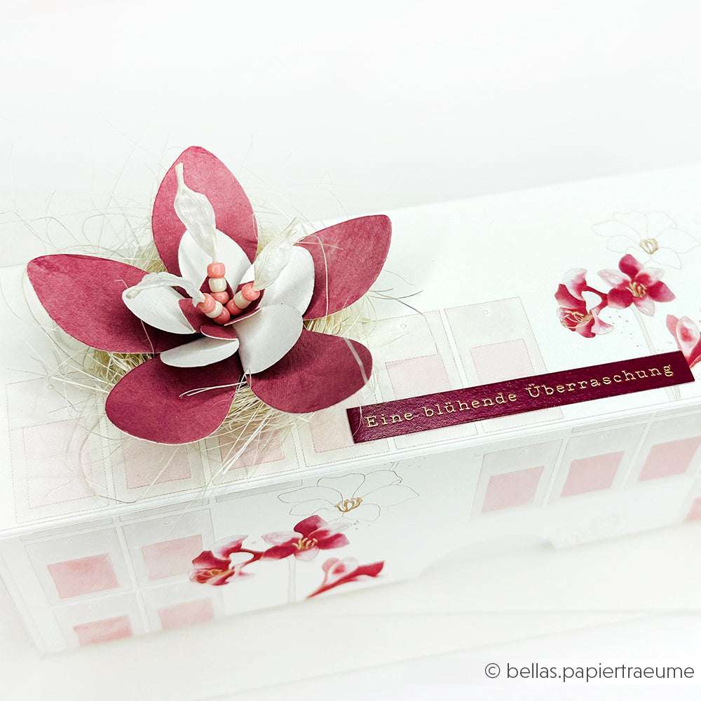 Rechteckige Schachtel von Alexandra Renke mit rosafarbener und weißer Papierblume, deutsches Etikett "Eine blühende Überraschung", ideal zum Basteln von Karten oder zum Versenden bunter Herbstgrüße.