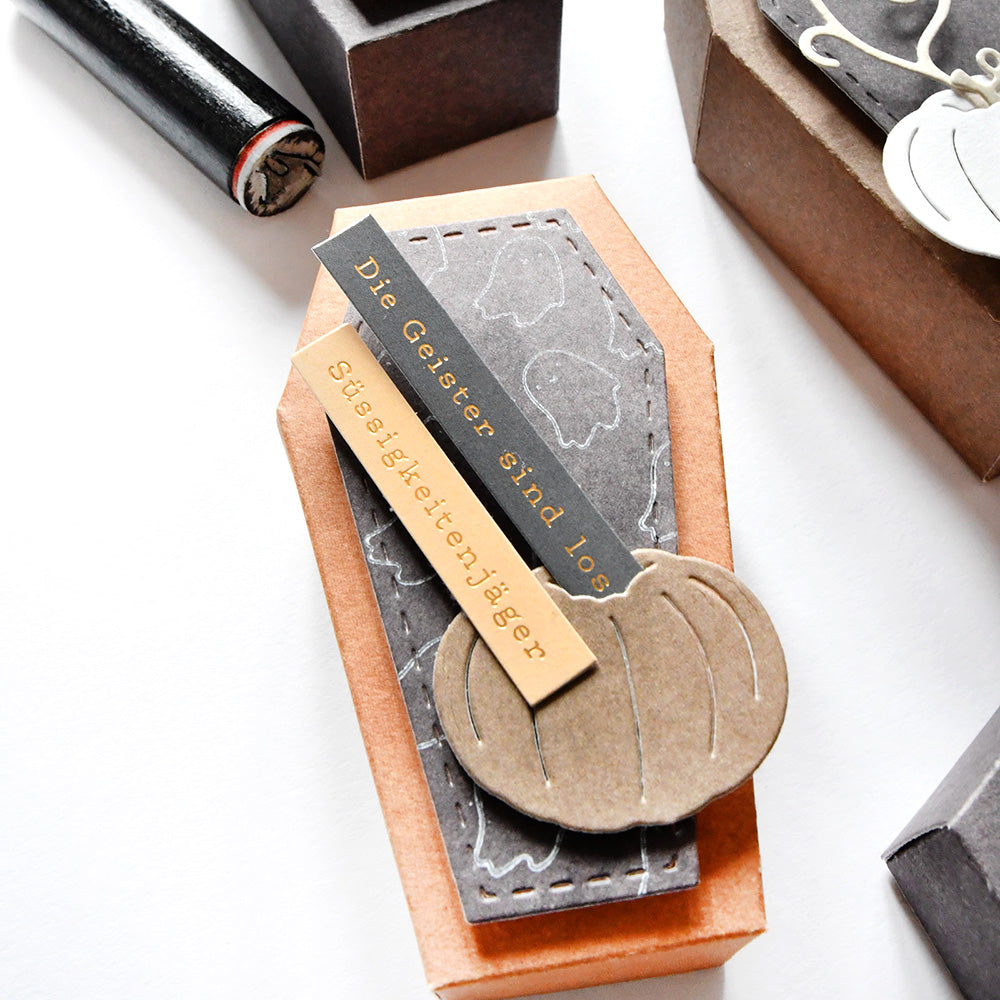 Alexandra Renke Halloween-Leckerbissen in Sargform mit deutschen Etiketten, gekrönt von einer Kürbisdekoration aus Papier und verziert mit Sticker-Streifen zur zusätzlichen Verschönerung.