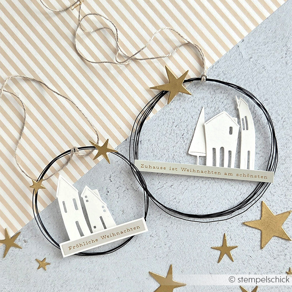 Zwei Alexandra Renke Drahtkränze mit weißen Häusern, goldenen Sternen und deutschen Weihnachtsgrüßen auf einem grauen, mit Sticker-Streifen versehenen Hintergrund verleihen Ihrer Dekoration den Charme der Weihnachtszeit.