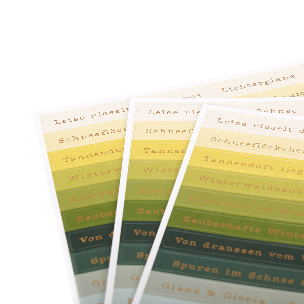 Drei Bögen Alexandra Renke Sticker-Streifen mit grünem und gelbem Farbverlauf und deutschem Text, aufgefächert auf weißem Hintergrund - ideal zum Basteln von Karten oder um Winterwaldzauber in Ihre kreativen Projekte zu bringen.