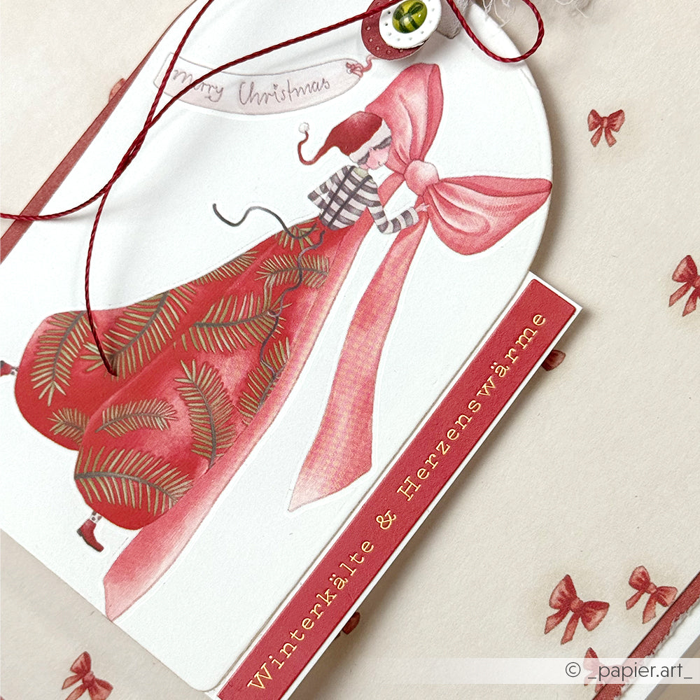 Geschenkanhänger von Alexandra Renke mit einer Illustration einer Frau in Rot, die eine rosa Schleife hält und dem Text "Winterkälte & Herzenswärme". Ideal für Ihre winterlichen Karten und kuscheligen Jahreszeitengrüße.