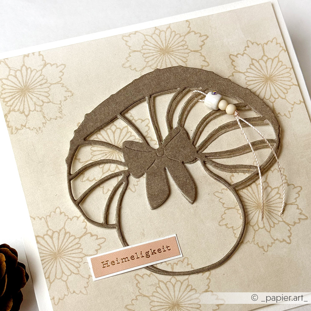 Handgemachte Karte von Alexandra Renke mit ausgeschnittenem Pilz, Schleife, Perlen und "Heimeligkeit" auf geblümtem Papier - ideal für die Kartengestaltung und um Wohlfühlweihnachtsflair in Ihre Urlaubsgrüße zu bringen.