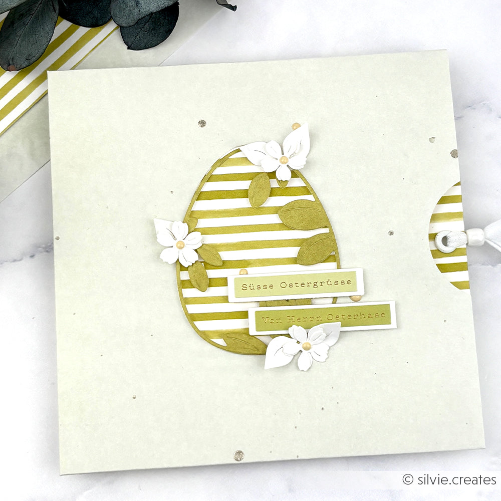 Eine handgemachte Osterkarte von Alexandra Renke mit Kartengestaltung mit Sticker-Streifen, einem gestreiften Ei, weißen Blumen und deutschem Text auf hellem Hintergrund - perfekt für die Osterzeit.