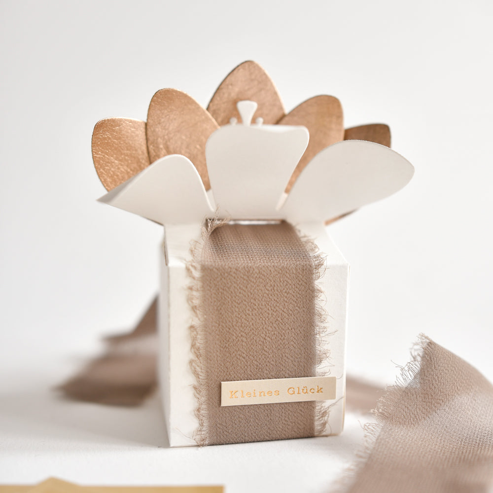 Kleine weiße Geschenkbox von Alexandra Renke mit einem beigen Stoffband und einem blumenförmigen Deckel mit der Aufschrift „Kleines Glück“ – eine bezaubernde Art, Momente des Glücks festzuhalten. Ideal zum Verpacken kleiner Geschenke, für kreative Bastelideen oder elegante Aufkleberstreifen.