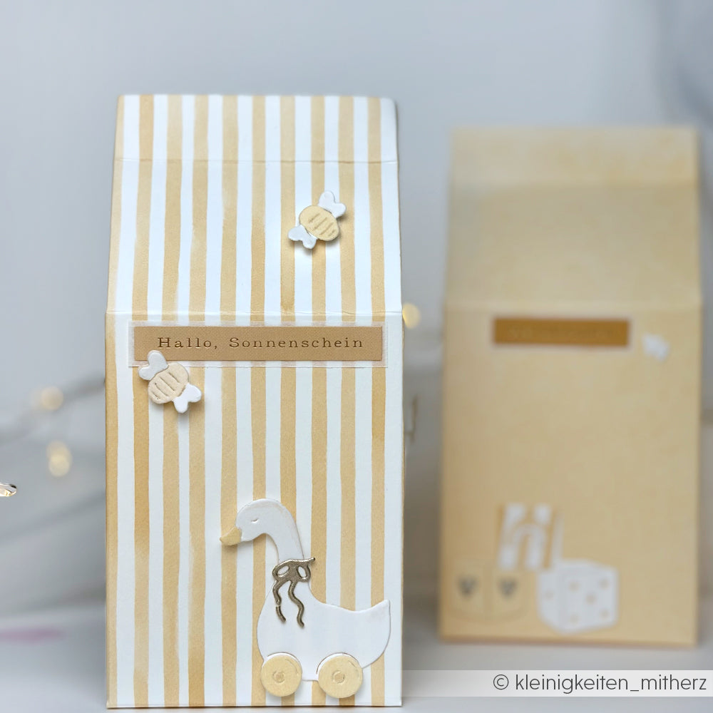 Gestreifte Geschenkbox von Alexandra Renke mit Sticker-Streifen, niedlichen Enten- und Bienenmotiven und "Hallo, Sonnenschein"-Aufkleber. Noch eine unscharfe Schachtel dahinter - perfekte Kartengestaltung für "Willkommen im Leben"!.