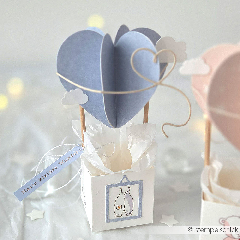 Ein blauer herzförmiger Papier-Heißluftballon von Alexandra Renke in einer weißen Schachtel mit Wolken, Sticker-Streifen-Akzenten und einem angebrachten "Willkommen im Leben"-Grußanhänger.