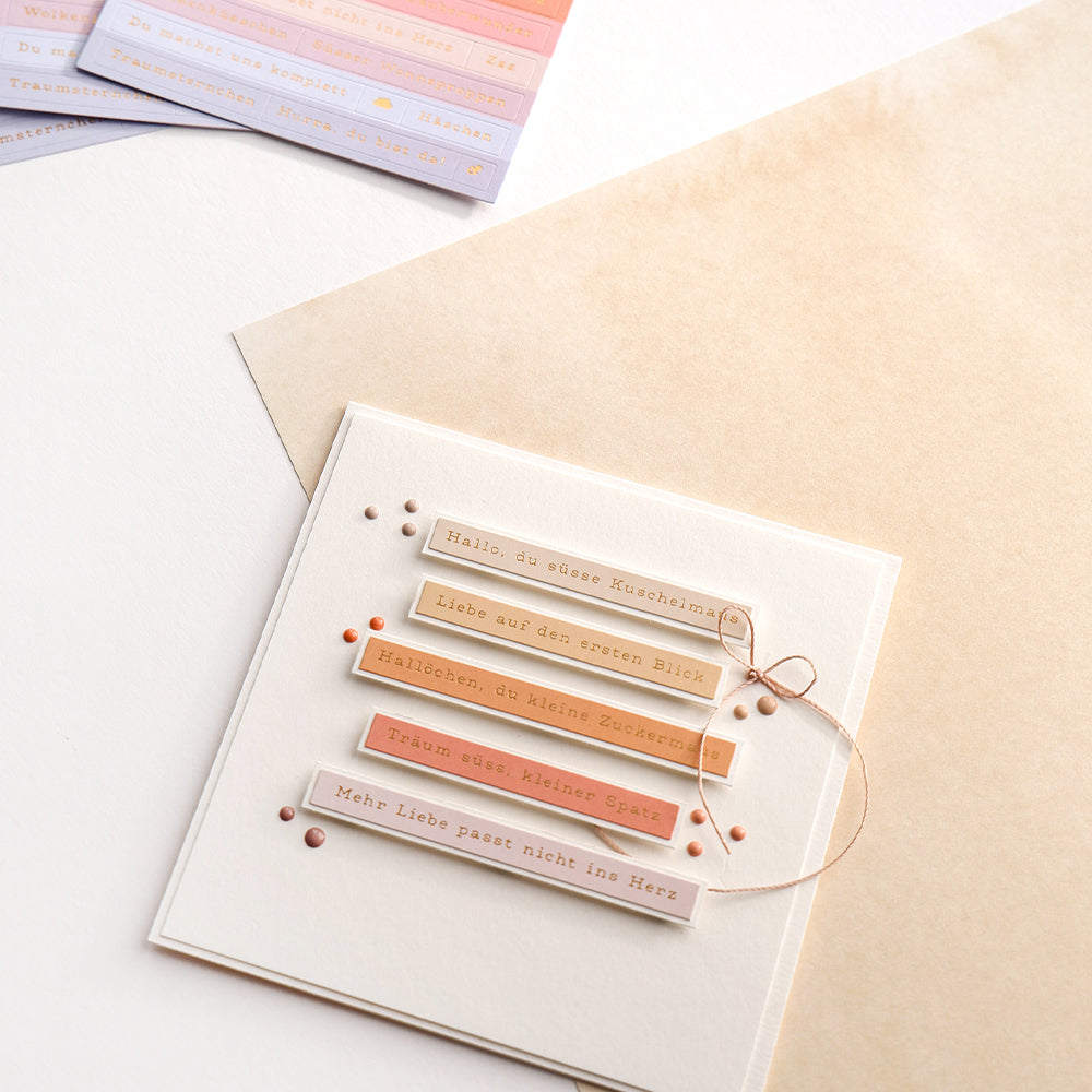 Eine handgefertigte Karte von Alexandra Renke mit deutschen Sprüchen, rosa und beigen Akzenten, Sticker-Streifen zur Dekoration und einer gebundenen Schnur auf neutralem Hintergrund.
