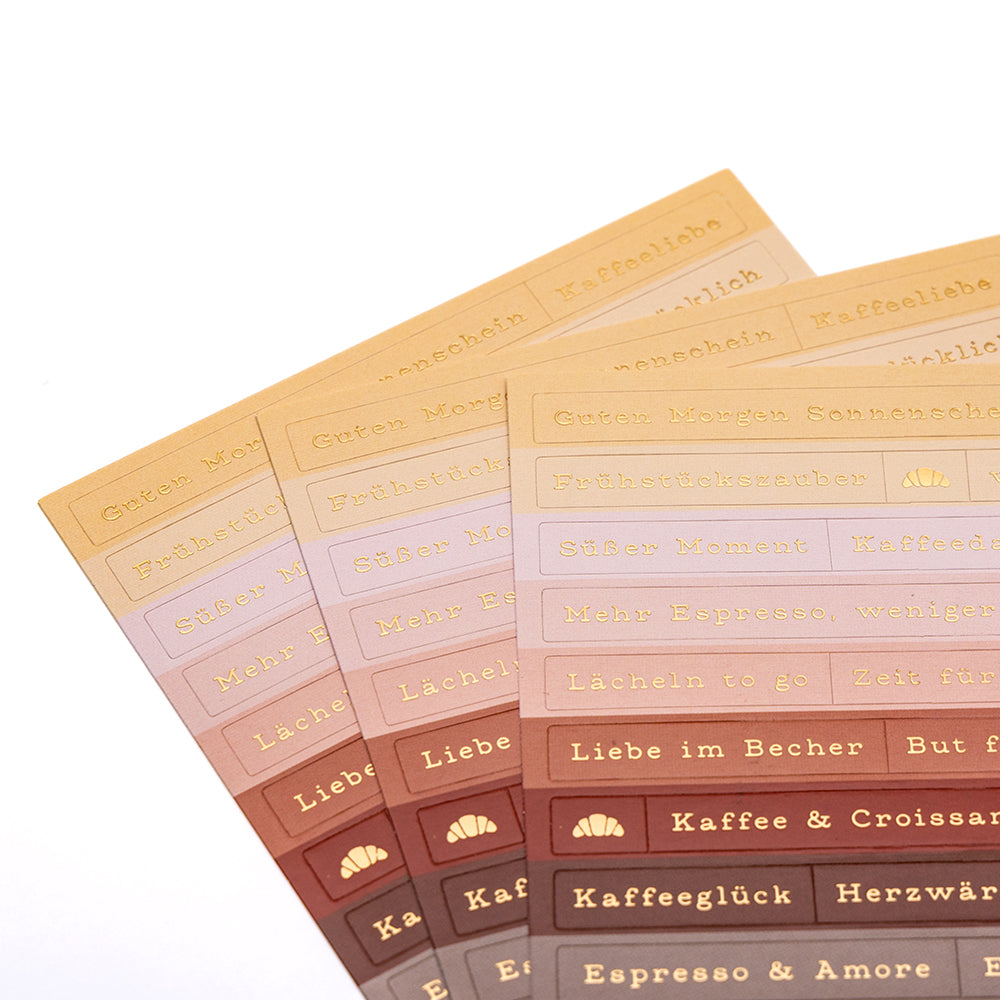 Vier Bögen mit bunten Alexandra Renke-Etiketten mit deutschem Text, Hörnchen-Symbolen und verspielten Sticker-Streifen, aufgefächert - ideal für die Kartengestaltung oder um Ihren Projekten einen Hauch von Kaffeezauber zu verleihen.