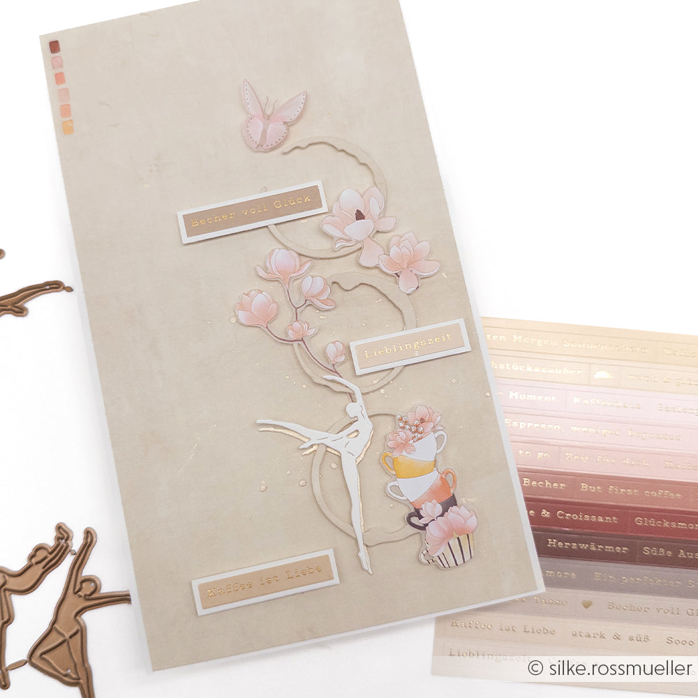 Eine beigefarbene Karte von Alexandra Renke mit rosa Blumen, Tassen und deutschem Text eignet sich perfekt für die Kartengestaltung. In Kombination mit metallischen Sticker-Streifen und Stanzteilen ist sie ideal, um Ihren Bastelprojekten einen Hauch von Kaffezauber zu verleihen.