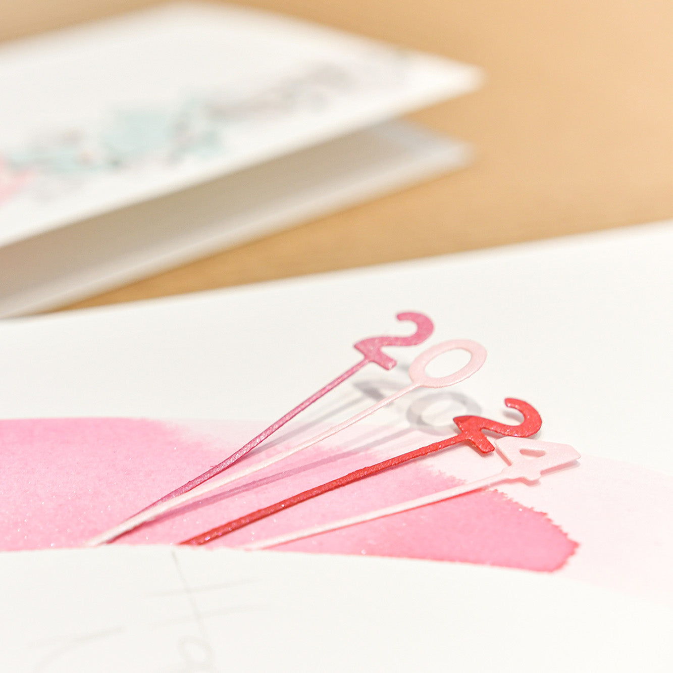 Nahaufnahme von rosa und roten 2024 Tortenauflegern, die mit Alexandra Renke Motivstanze Zahlen am Stiel Dies auf einer weißen und rosa Designpapierkarte hergestellt wurden, mit einer unscharfen Karte im Hintergrund.