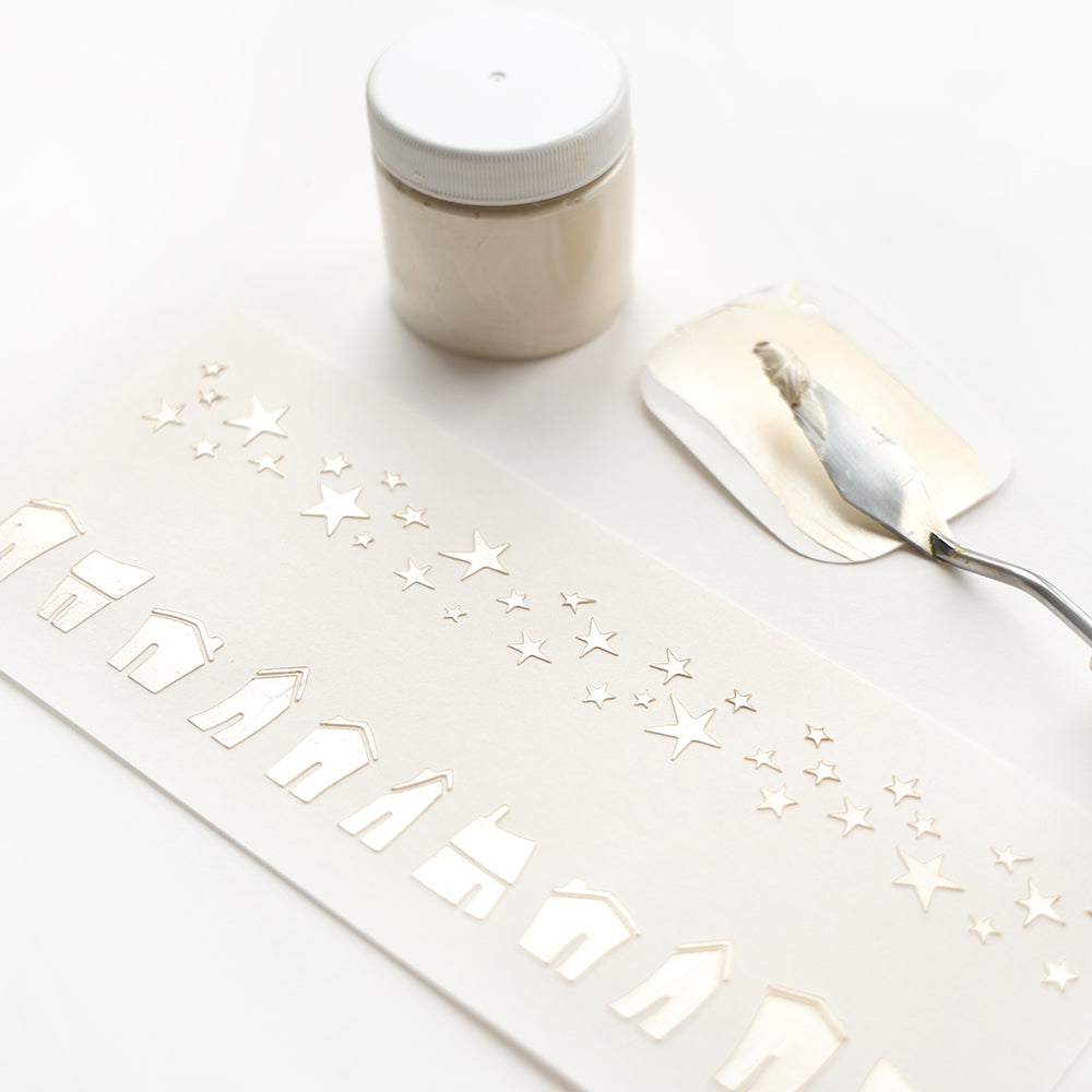 Alexandra Renke weiße Schablone mit Häusern und Sternen, Spachtel und Tiegel mit weißer Paste auf weißem Untergrund - ideal für kreative Schablonen und Schablonenprojekte.