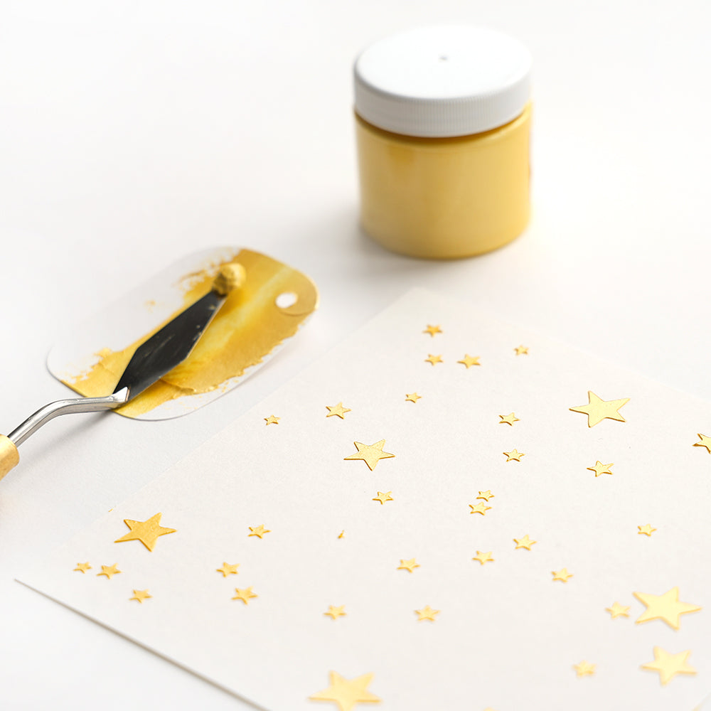 Alexandra Renke's Palettenmesser, gelbe Farbe, sternförmige Schablonen auf Papier und ein Glas gelber Farbe auf weißem Untergrund ergeben ein wiederverwendbares Stencil Sternenhimmel-Design.