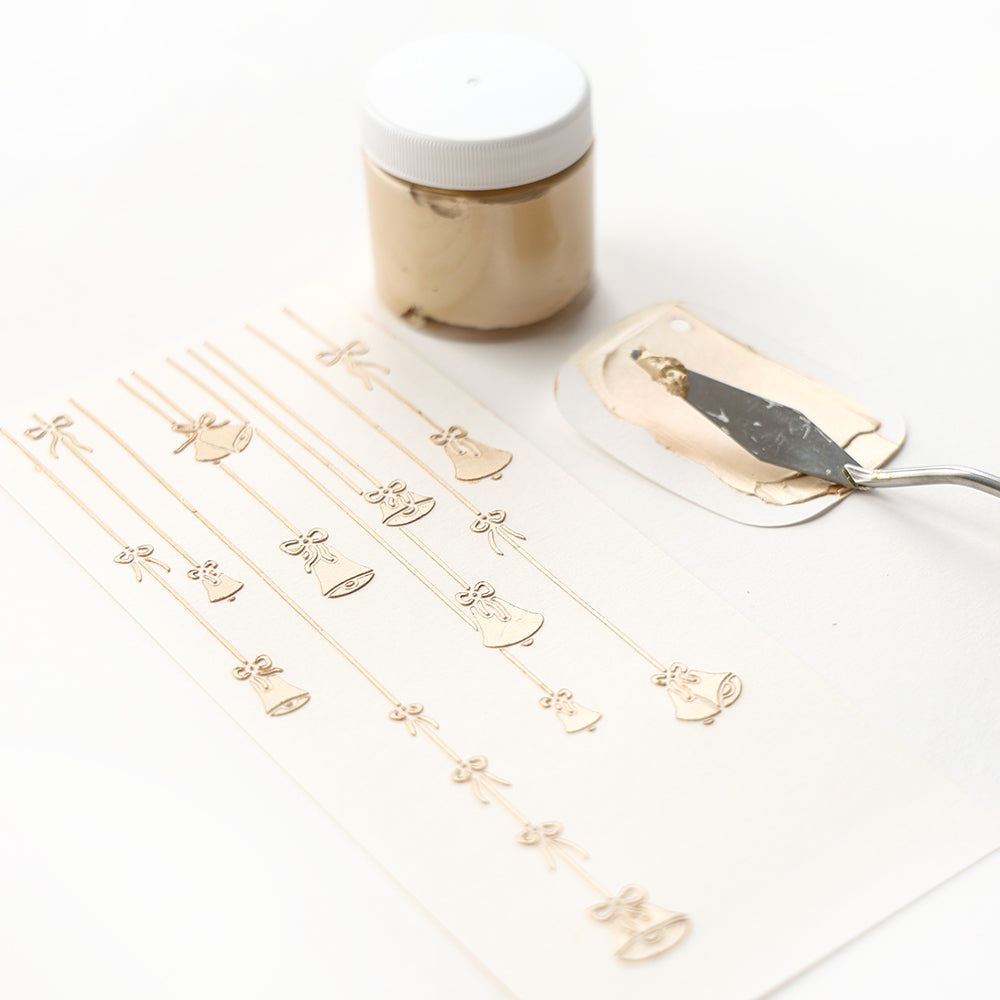 Goldglockenmotive auf Papier, hergestellt mit der Alexandra Renke Schablone Klingende Glocken und wiederverwendbaren Schablonen, abgebildet neben einem Goldpastendöschen und Spachtel. Ideal für Schablonen Papierprojekte.