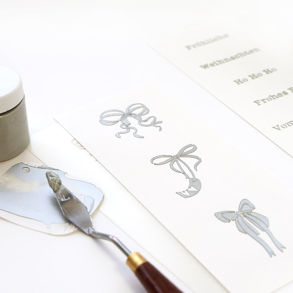 Alexandra Renke zeigt graue Farbe und einen Spachtel mit weißem Papier, einer Schablone und Zauberhaftes Schleifen-Trio Silberbogen-Illustrationen neben gedrucktem Text auf einer weißen Fläche.