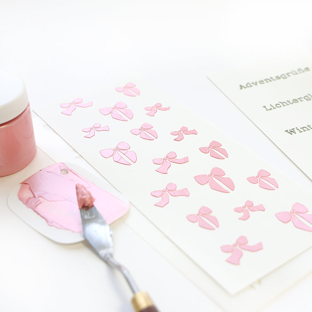 Gestalten Sie rosa Schleifen auf weißem Papier mit der Alexandra Renke Schleifenregen-Schablone, einem Spachtel, rosa Farbe und einem Farbtopf in der Nähe.