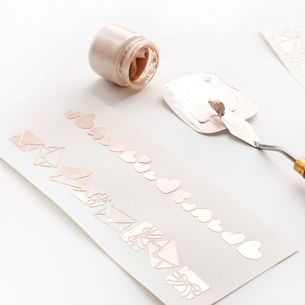 Die Papierstreifen von Alexandra Renke zeigen schablonierte Herzen und Umschläge in Metallicrosa, die mit wiederverwendbaren Schablonen hergestellt wurden, neben einem offenen Glas mit passender Farbe und einem Spachtel.