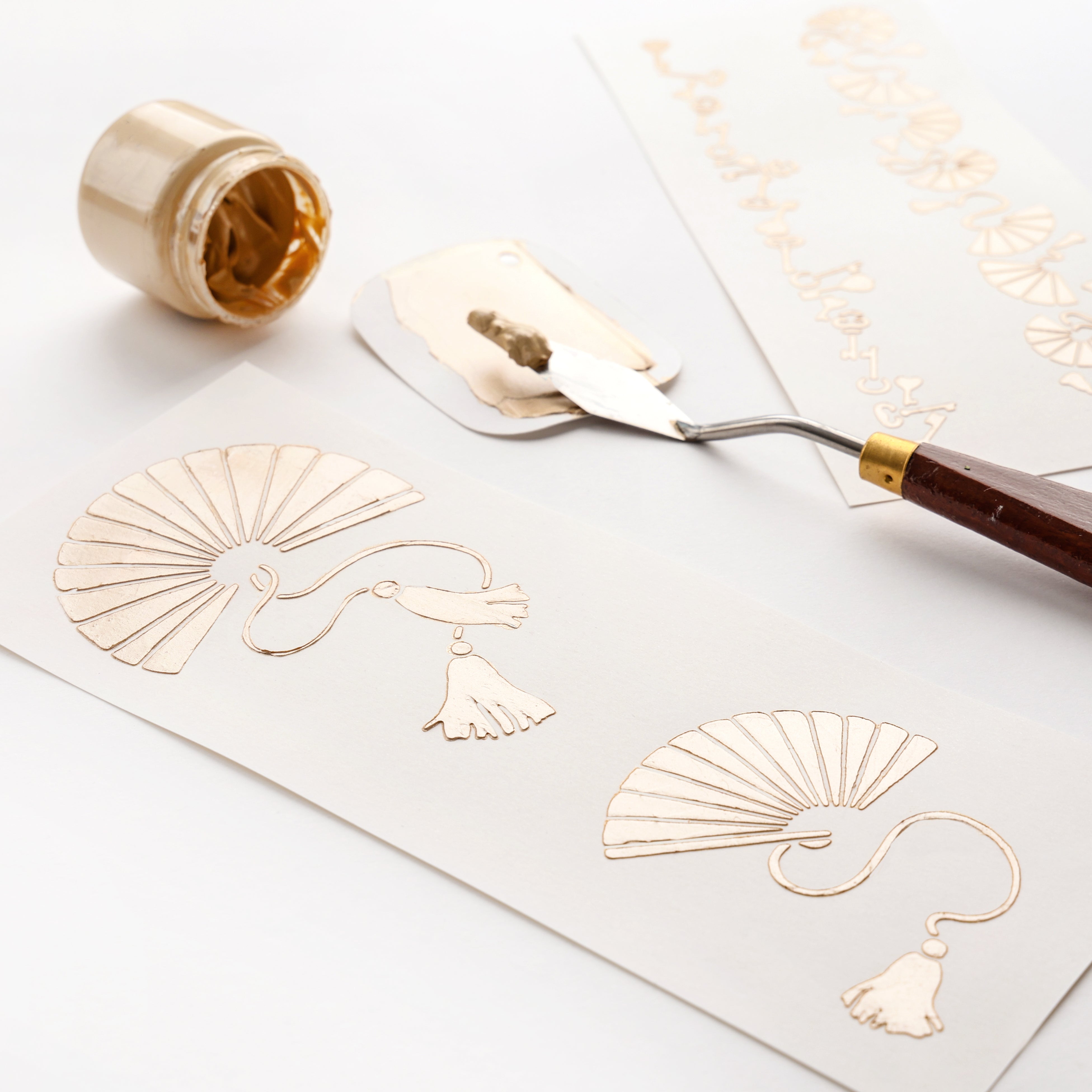 Goldfarbe und Spachtel neben eleganten Fächer- und Quastenmustern auf weißem Papier, erstellt mit dem Stencil Fächer-ideal von Alexandra Renke für Schablonen Papierprojekte.