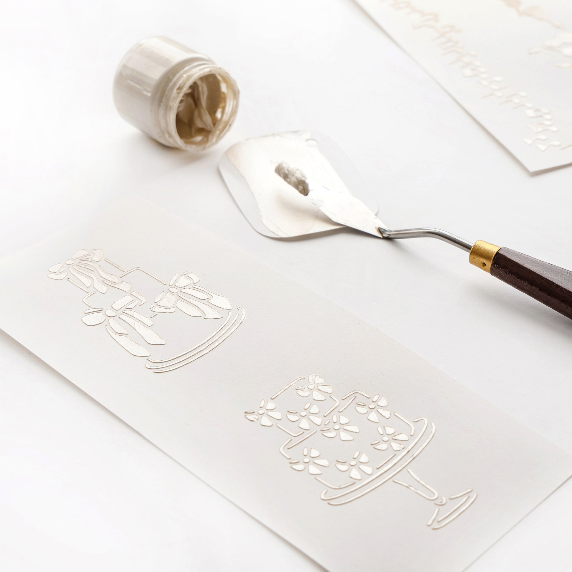 Mit einem Spachtel wird weißer Kleister mit wiederverwendbaren Schablonen von Alexandra Renke über geprägte Tortenmotive auf Papier aufgetragen. Ein offenes Glas in der Nähe unterstreicht die kreative Vielseitigkeit dieser Werkzeuge.