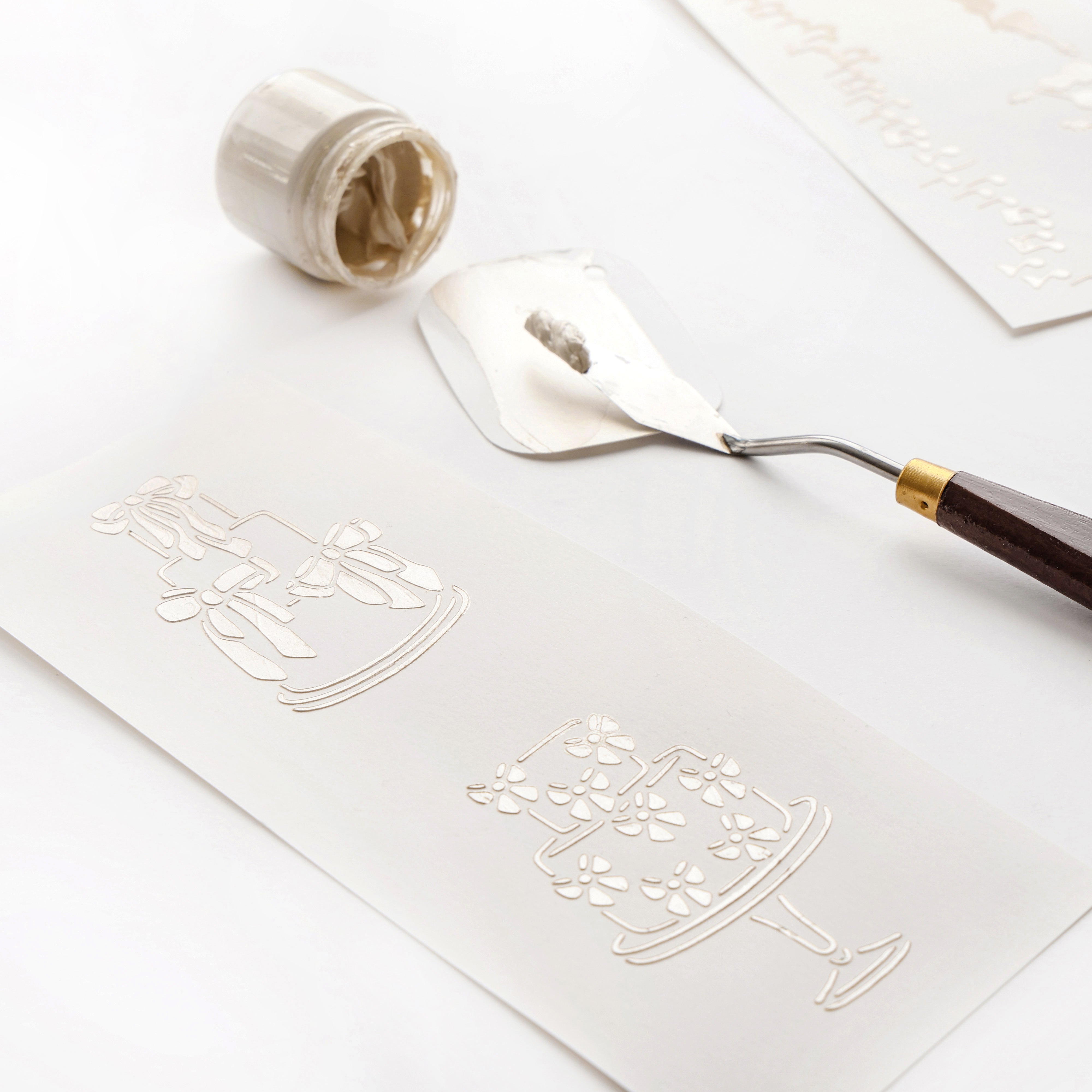 Mit einem Spachtel wird weißer Kleister mit wiederverwendbaren Schablonen von Alexandra Renke über geprägte Tortenmotive auf Papier aufgetragen. Ein offenes Glas in der Nähe unterstreicht die kreative Vielseitigkeit dieser Werkzeuge.