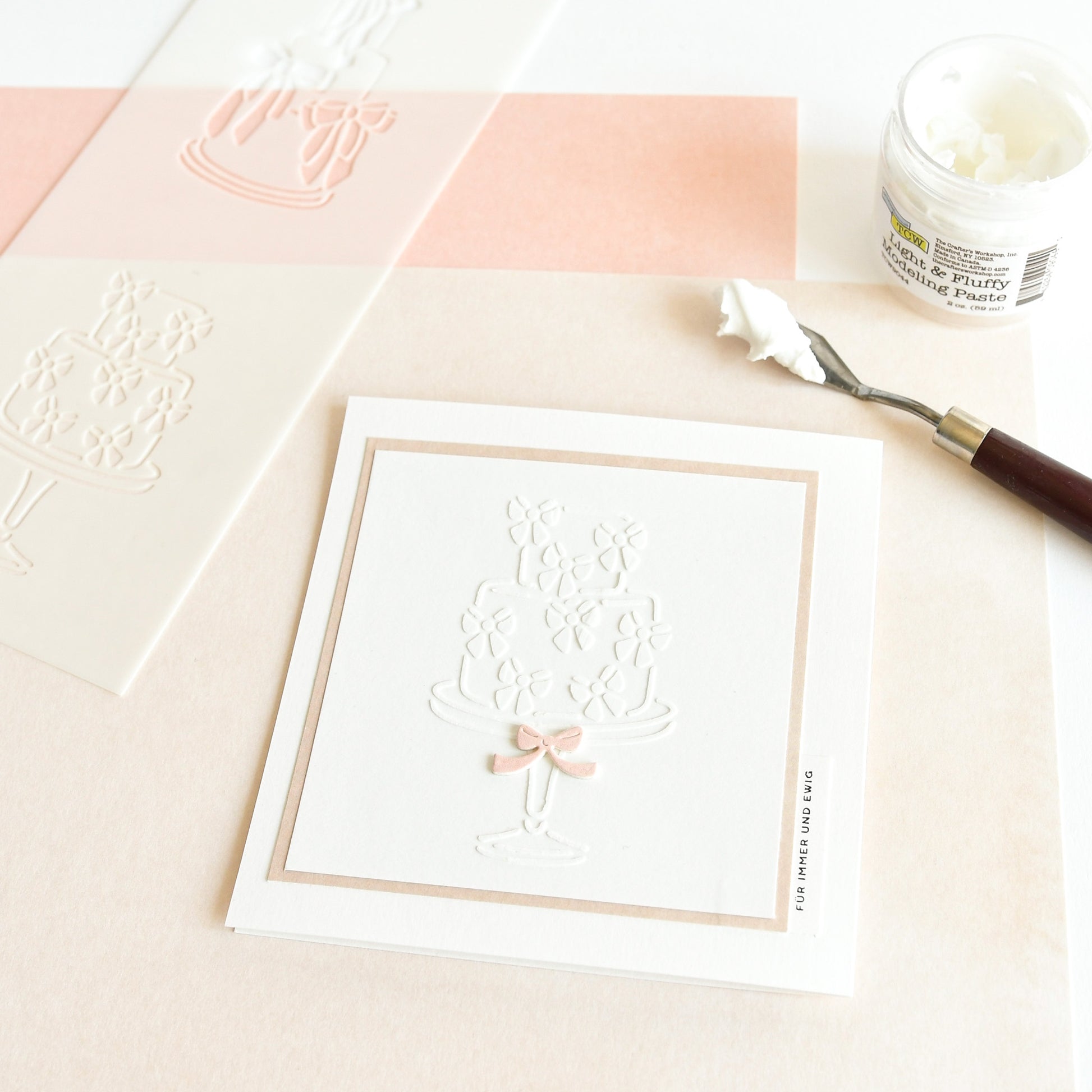 Quadratische Karte mit geprägtem Tortenmotiv, rosa Schleife und Bastelmaterial auf pfirsichfarbener Oberfläche - perfekt für Schablonen für Papierprojekte oder mit Ihrer Lieblingsschablone für wiederverwendbare Schablonen von Alexandra Renke für kreatives Flair.