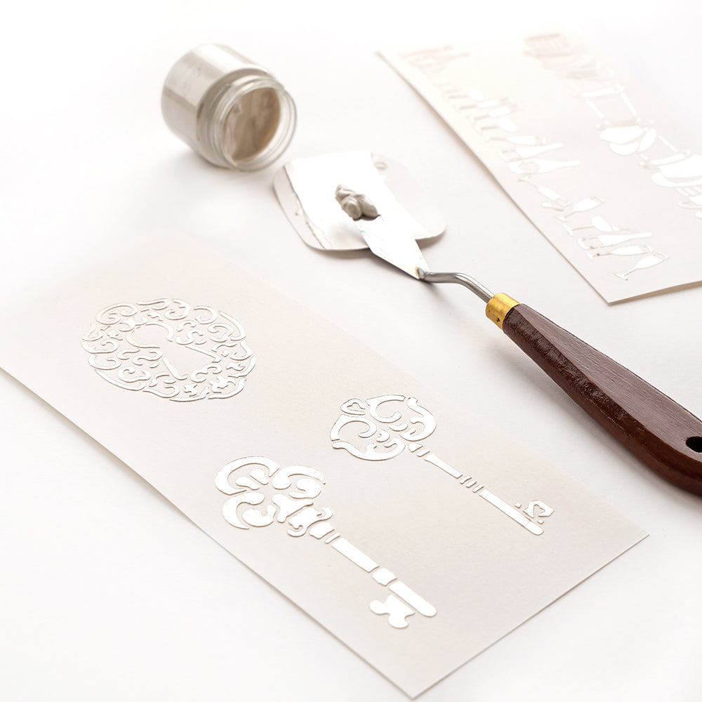 Ein Spachtel mit weißem Kleister, wiederverwendbare Schablonen und elegante Alexandra Renke Stencil Liebliche Schlüssel Designs auf weißem Papier.