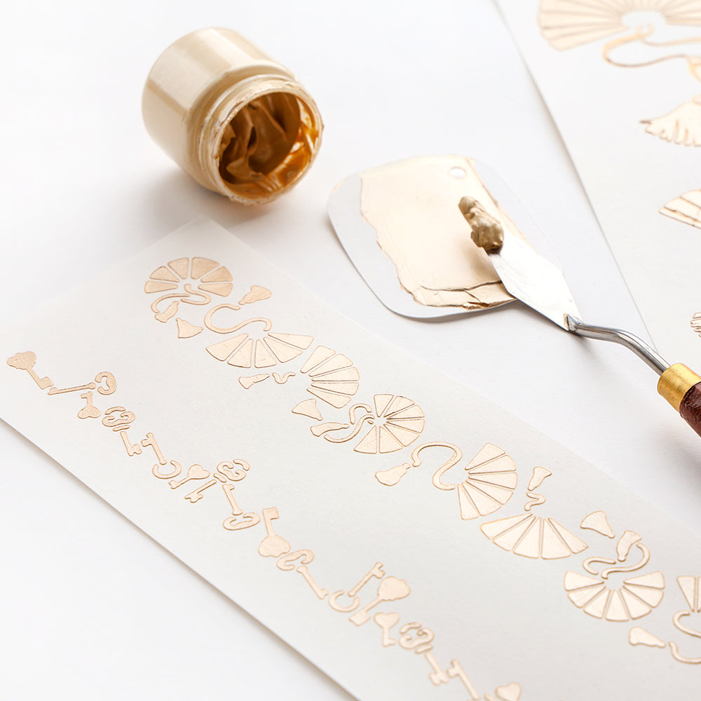 Alexandra Renke Goldfarbe und ein Spachtel stehen neben Papier mit dekorativen Goldmustern und Schablonen-Musterstreifen, die für wiederholte kreative Projekte wiederverwendet werden können.