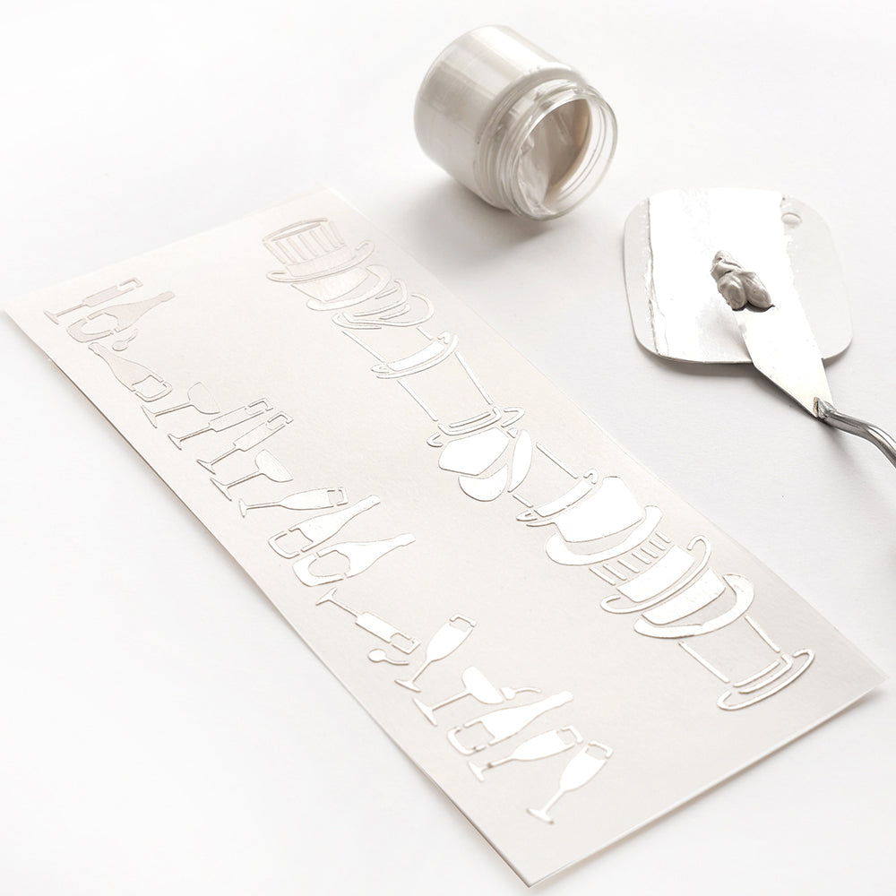 Alexandra Renke weiße Strukturpaste wird auf eine wiederverwendbare Schablone Musterstreifen auf Papier aufgetragen, wobei ein offenes Glas und ein Spachtel neben dem Projekt stehen.