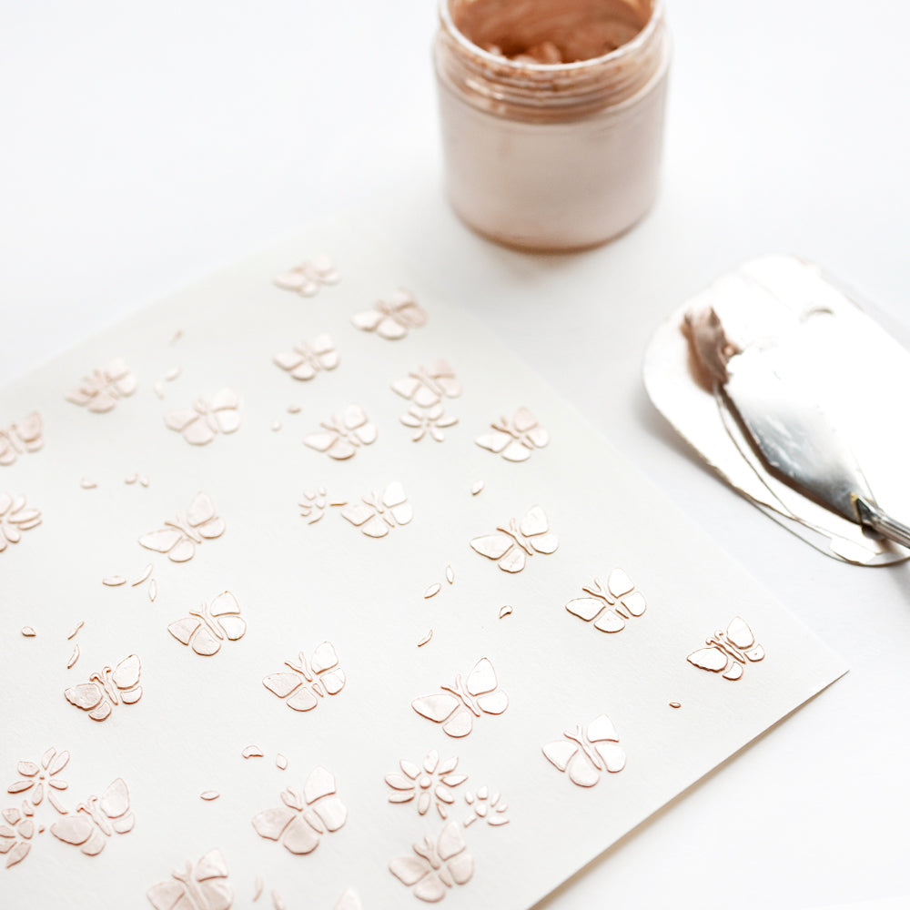 Eine weiße Alexandra Renke Schablonen Papierprojekte mit geprägter roségoldener Schablone Florale Schmetterlinge steht neben einem offenen Glas mit Metallpaste und einem Spachtel.