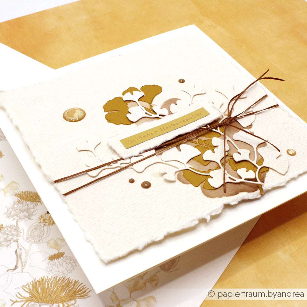 Quadratische, handgefertigte Karte mit goldenem Blumenmuster, brauner Schnur und dekorativen Aussparungen auf weißem Strukturpapier, hergestellt mit der wiederverwendbaren Schablonenschablone Ginkgowirbel von Alexandra Renke für zusätzliche Eleganz.