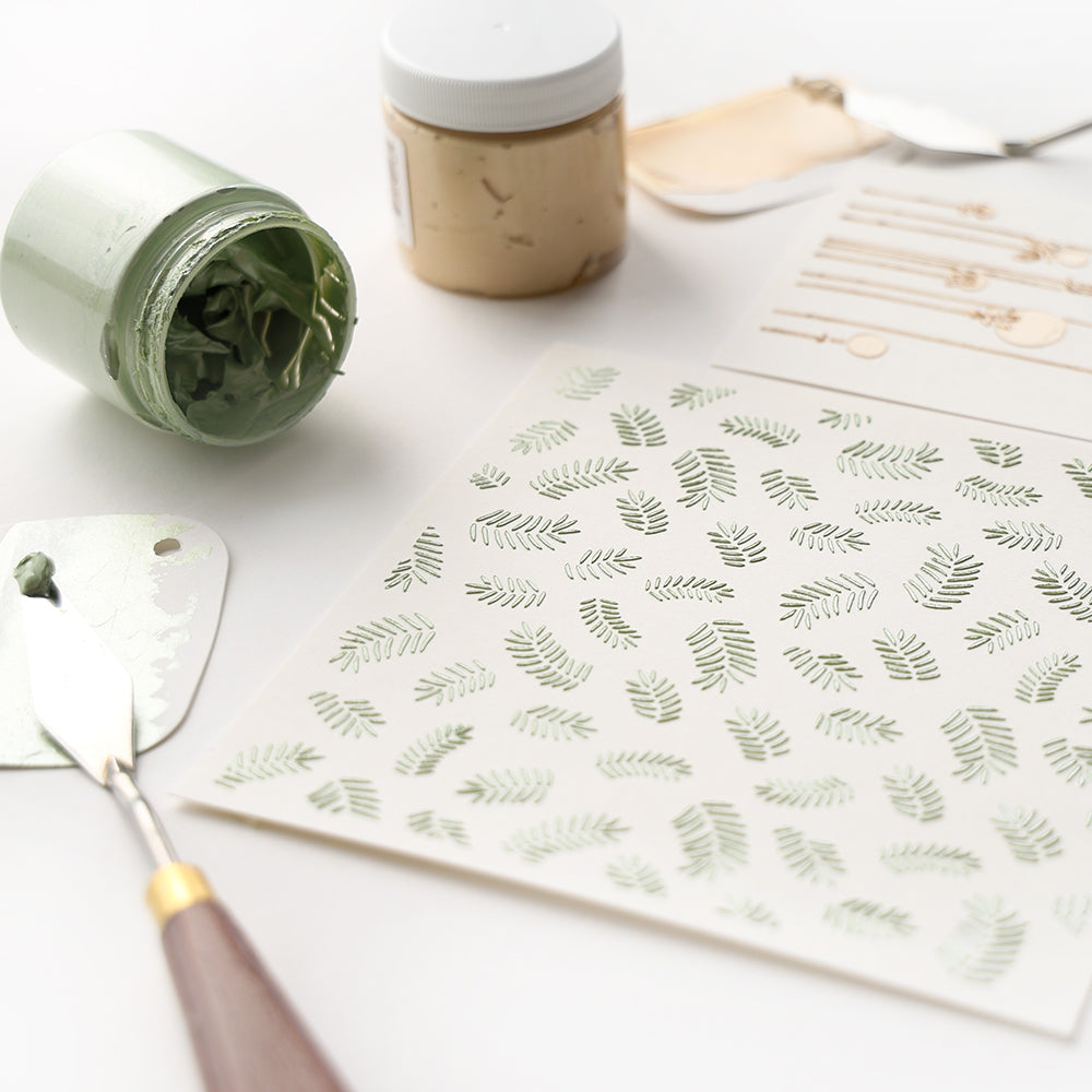 Palettenmesser, Farbdosen und wiederverwendbare Alexandra Renke Schablonen mit grünen Blattmustern, darunter die Schablone Tannenzweige, angeordnet auf einer weißen Fläche.
