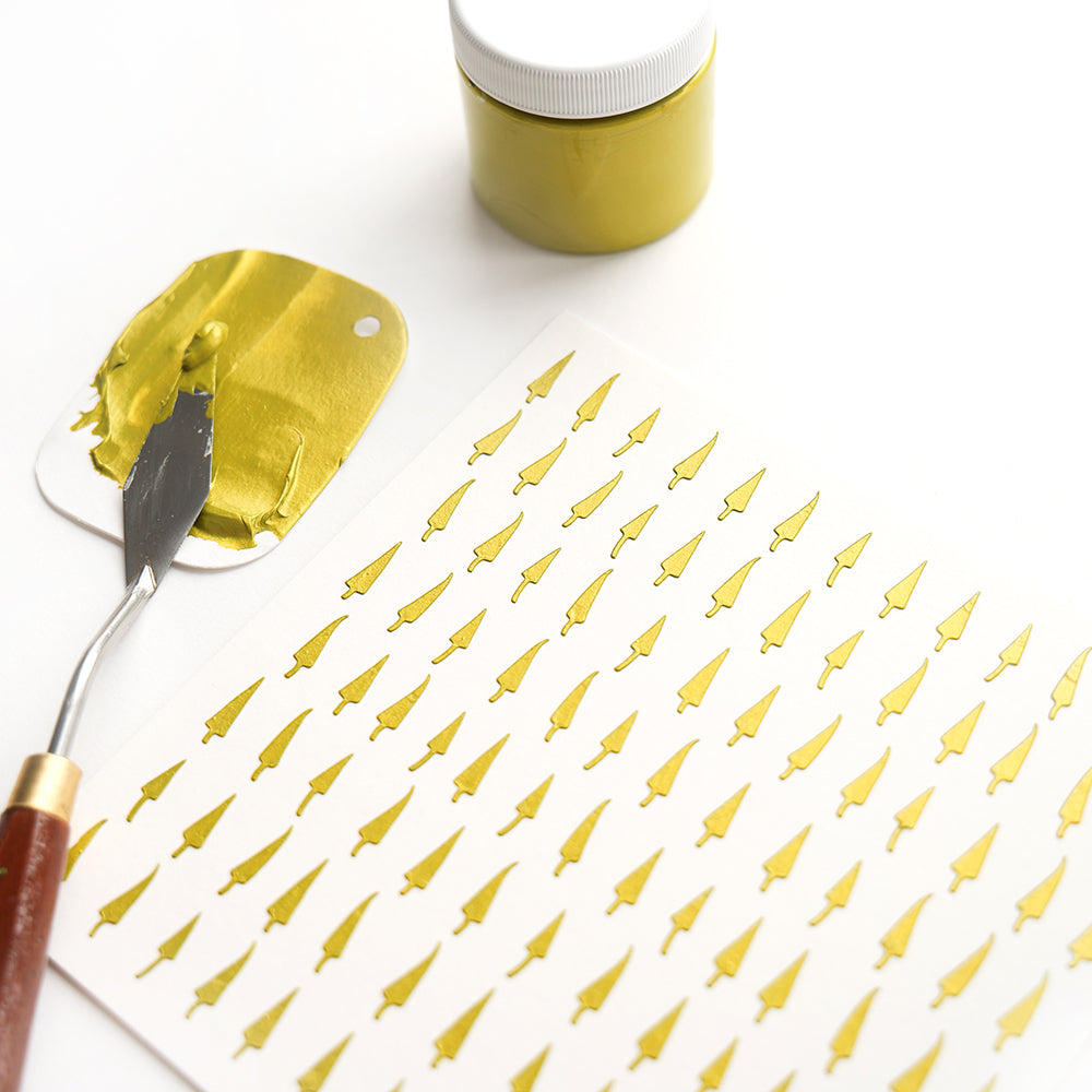 Ein Alexandra Renke Spachtel mit goldener Farbe, ein goldener Farbtopf, wiederverwendbare Schablonen und Papier mit Schablonenkunst und goldenen Pinselmustern werden auf weißem Hintergrund gezeigt.