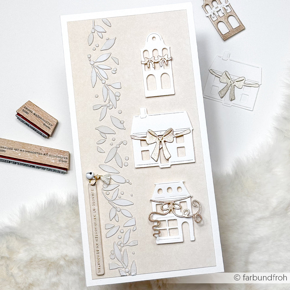 Handgemachte Karte von Alexandra Renke mit gestanzten weißen Häusern, Schleifen und belaubten Zweigen auf beigem Hintergrund. Enthält wiederverwendbare Schablonen und Stencil Musterstreifen Beeren Akzente mit Gummistempeln in der Nähe.
