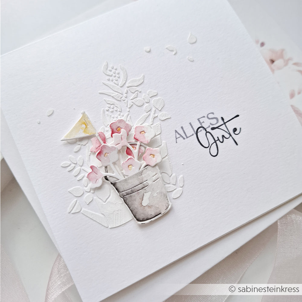 Grußkarte von Alexandra Renke mit geprägten Blumen, einem grauen Eimer voller rosa Blüten und dem deutschen Schriftzug „ALLES Gute“ auf weißem Hintergrund – ideal zum Basteln mit einer wiederverwendbaren Schablone oder der Schablone „Gartenwirbel“.