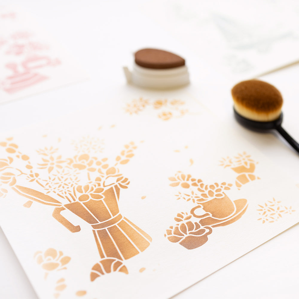 Schablonenkunst von Blumen in einer Gießkanne mit Alexandra Renke Schablonen und braunen Stempelkissen, mit Pinseln auf einer weißen Fläche. Die wiederverwendbaren Schablonen verleihen diesem einzigartigen Design einen kreativen Charme.