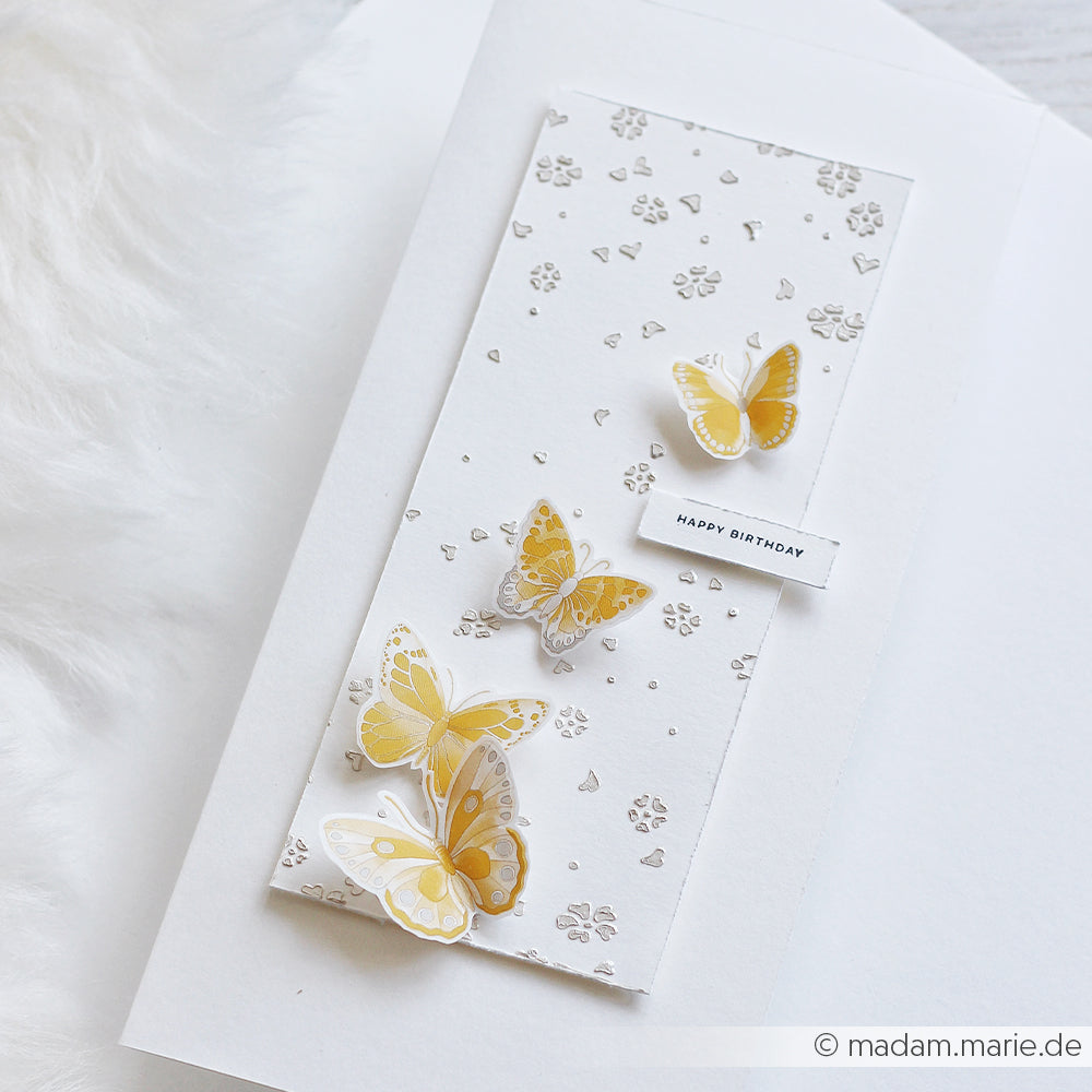 Handgefertigte Geburtstagskarte mit drei gelben Schmetterlingen und einem „Happy Birthday“-Schriftzug auf strukturiertem weißem Karton, gestaltet mit der wiederverwendbaren Schablone „Herzblütenfall“ von Alexandra Renke für den besonderen Charme.