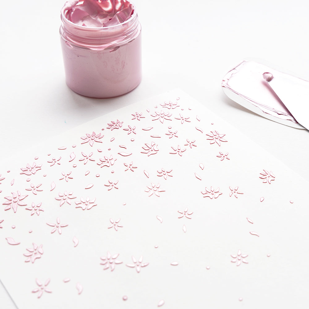 Ein weißes Papier mit rosa geprägten Blumenmustern, ein Glas mit rosa Kleister und ein Spachtel - ideal zum Basteln mit Alexandra Renke transparente wiederverwendbare Schablonen Schablonen.