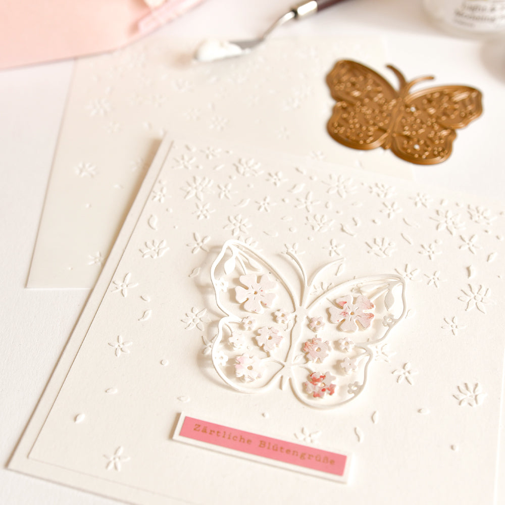 Die weiße Grußkarte von Alexandra Renke besticht durch einen Schmetterlingsausschnitt und geprägte Blumen, kombiniert mit einem goldfarbenen Schmetterlingsstanzteil – ideal für Schablonen-Papierprojekte oder die Verwendung von wiederverwendbaren Schablonen wie „Stencil Sternblütenfall“.