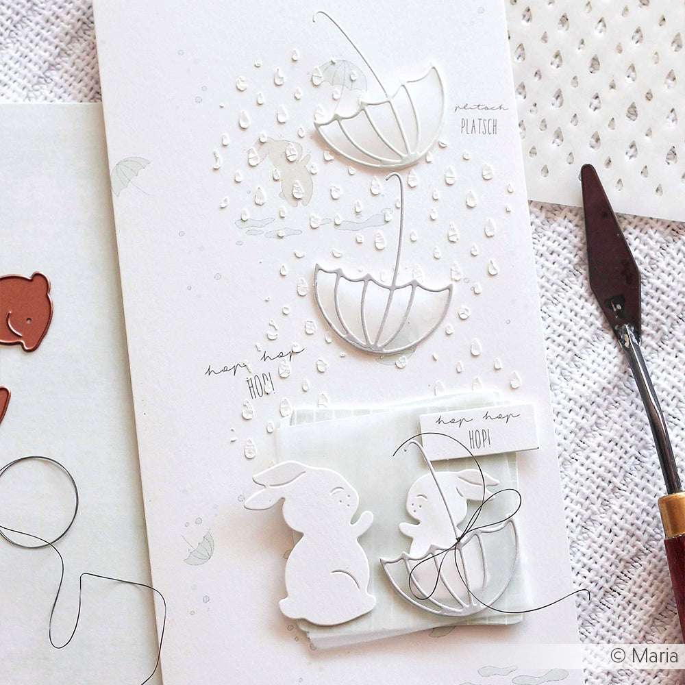 Zwei weiße Papierhasen mit Regenschirmen erscheinen auf einer Karte, die mit geprägten Regentropfen unter Verwendung der Alexandra Renke Schablone Regentropfen und dezenten Hasenillustrationen verziert ist - eine charmante Idee für Papierbastelprojekte.