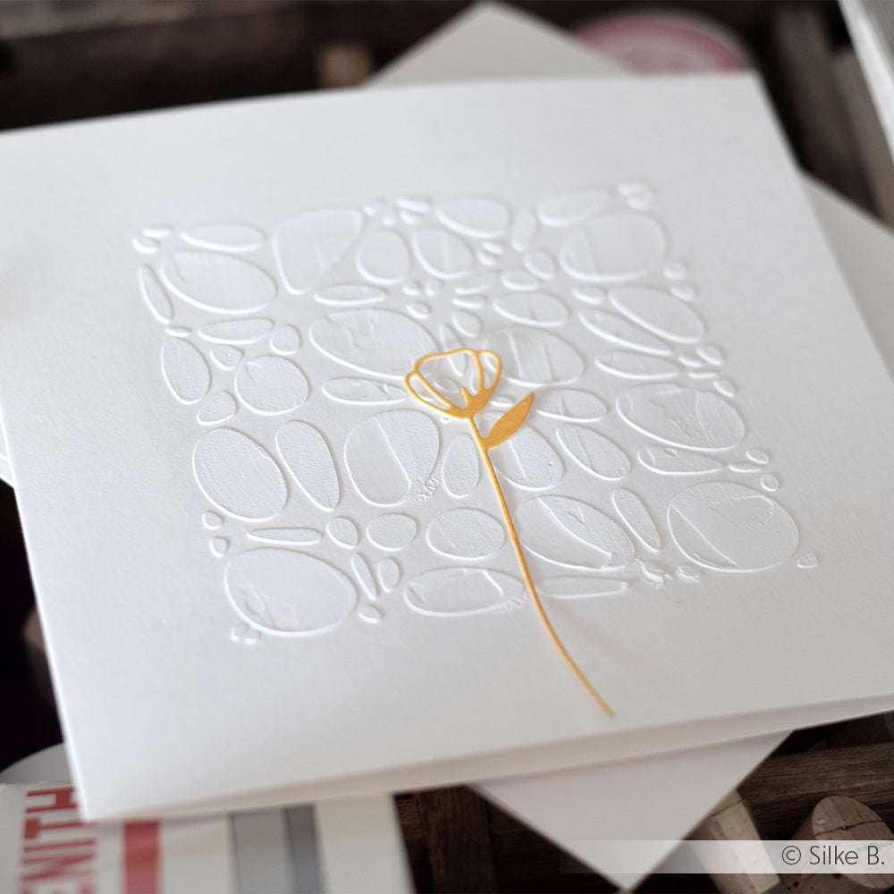 Weiße Karte mit geprägten Kieselsteinen und einem einzelnen gelben Blumenstängel in der Mitte, hergestellt mit den wiederverwendbaren Schablonen von Alexandra Renke - ideal für Schablonen-Papierprojekte und um jeder handgemachten Kreation eine einzigartige Textur zu verleihen.