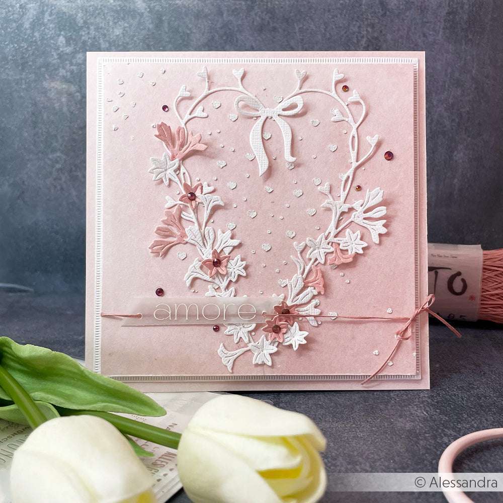 Eine rosafarbene Grußkarte von Alexandra Renke mit einem herzförmigen Blumenkranz, einer Schleife und "amore" auf der Vorderseite - ideal für Papierprojekte oder zur Verwendung mit Schablonen.