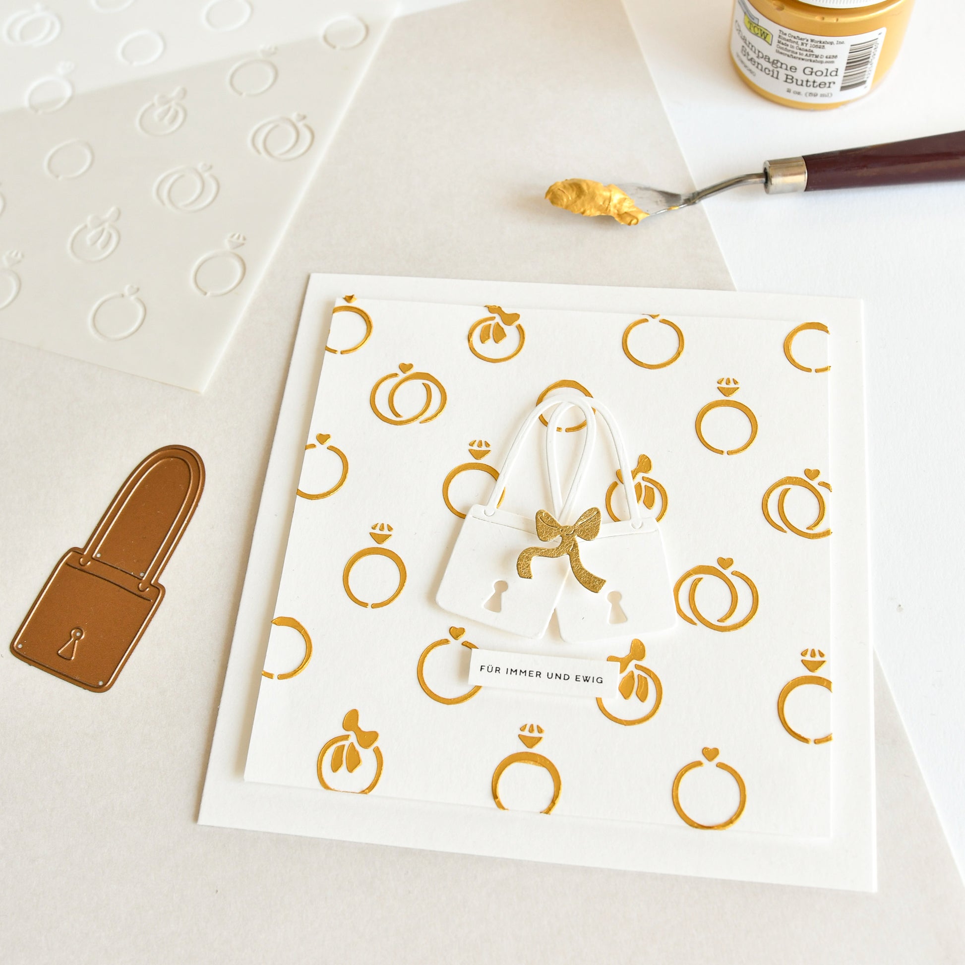 Weiße Karte mit goldenen Ring- und Schlossmustern, die mit Schablonen von Alexandra Renke hergestellt wurden; eine weiße Geldbörse mit goldener Schleife ist neben dem Kunstzubehör abgebildet.