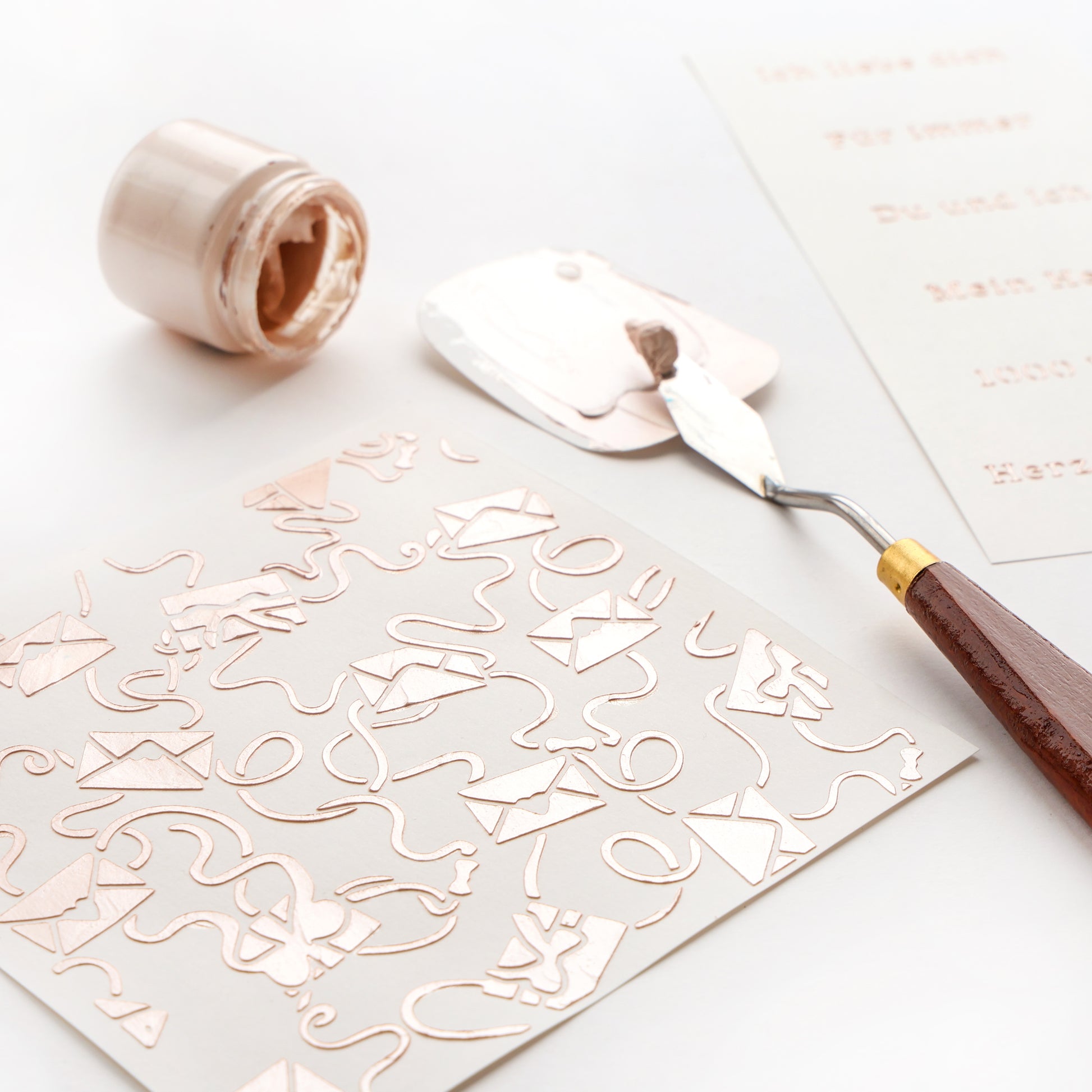 Mit einem Spachtel wird Metallic-Farbe auf eine Alexandra Renke-Schablone Verschlungene Liebesbriefe mit Umschlägen auf weißem Papier aufgetragen; ein nahe gelegener Farbtopf ist ideal für Schablonen Papierprojekte und wiederverwendbare Schablonen.