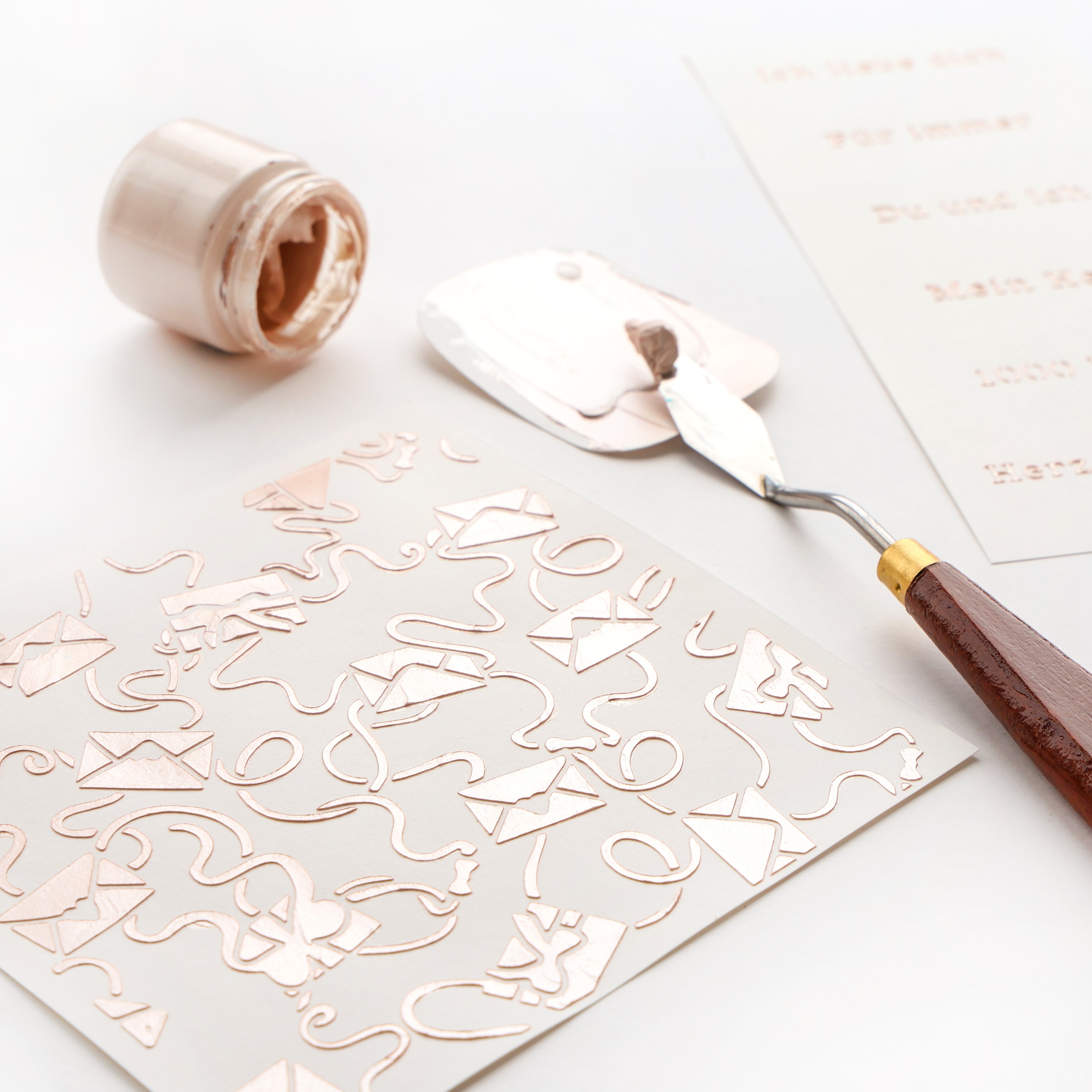 Mit einem Spachtel wird Metallic-Farbe auf eine Alexandra Renke-Schablone Verschlungene Liebesbriefe mit Umschlägen auf weißem Papier aufgetragen; ein nahe gelegener Farbtopf ist ideal für Schablonen Papierprojekte und wiederverwendbare Schablonen.