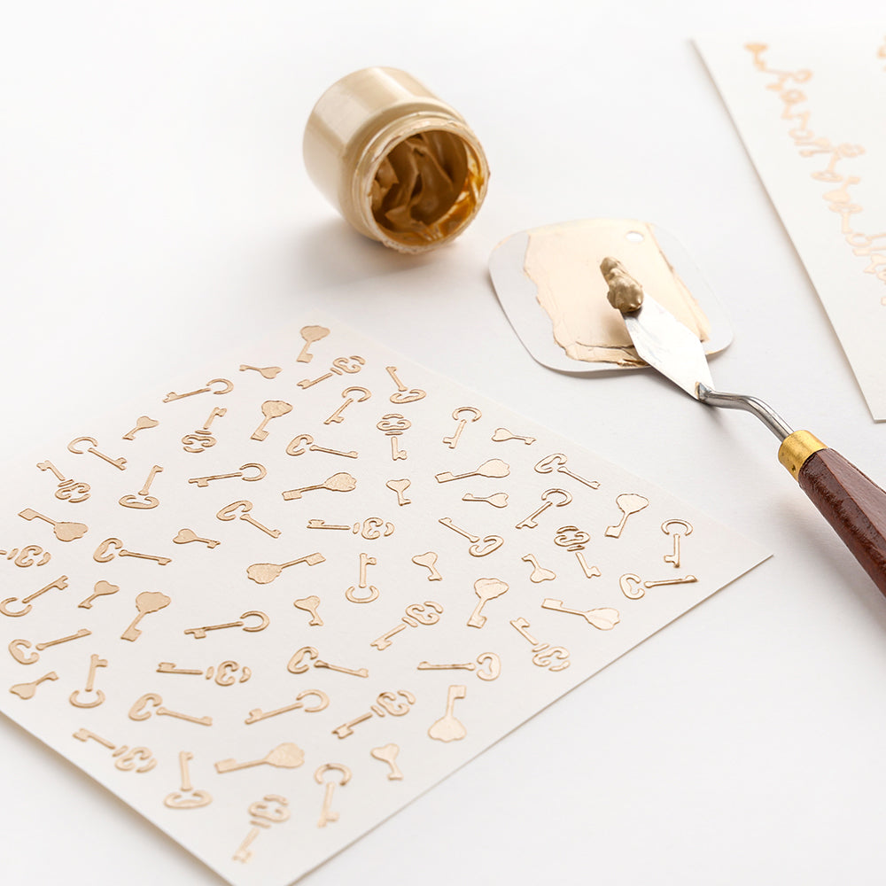 Goldene Schlüsselmotive auf weißem Papier, Schablone, Spachtel und ein offener goldener Farbtopf von Alexandra Renke sind auf einer weißen Fläche angeordnet - die perfekte Inspiration für Papierprojekte.