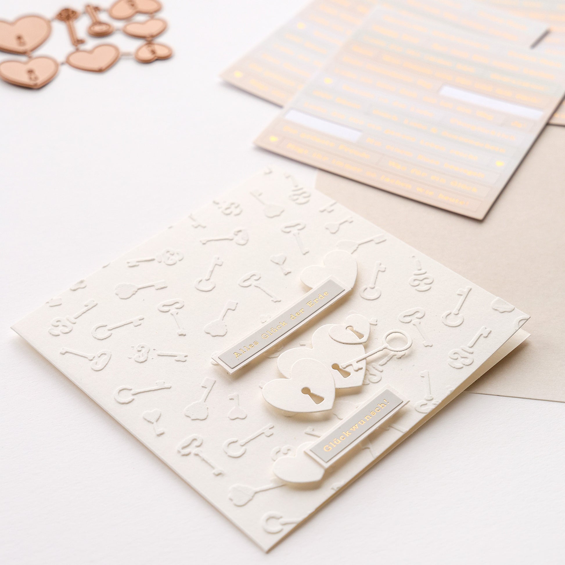 Cremefarbene Grußkarte von Alexandra Renke mit Herz- und Schlüsselmotiven, metallischen Herzanhängern, dekorativen Stickern und Details, die mit wiederverwendbaren Schablonen aus der Stencil Schlüsselzauber-Kollektion hergestellt wurden.