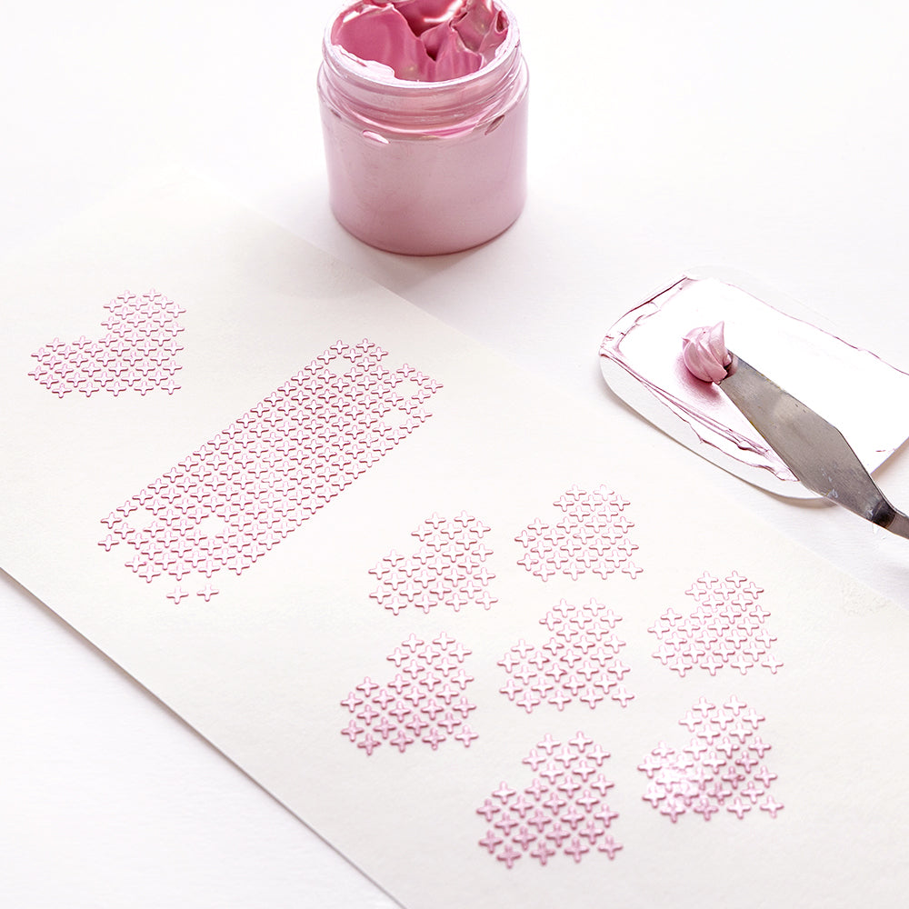 Rosa Schablone Herzen und Rechtecke auf weißem Papier, mit einem Glas rosa Kleister und Spachtel in der Nähe. Alexandra Renke wiederverwendbar Schablonen erstellen charmante Stencil Kreuzstich Herzmuster für vielseitige Handwerksprojekte.