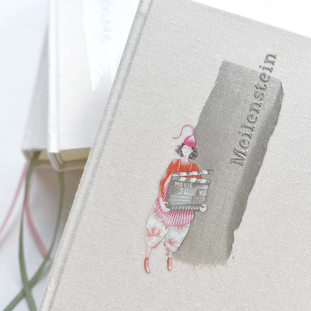 Das beigefarbene Notizbuch von Alexandra Renke mit der Illustration einer Person, die eine Filmklappe in der Hand hält, ist ideal für Papierprojekte oder das Skizzieren eigener Entwürfe mit Schablonen.