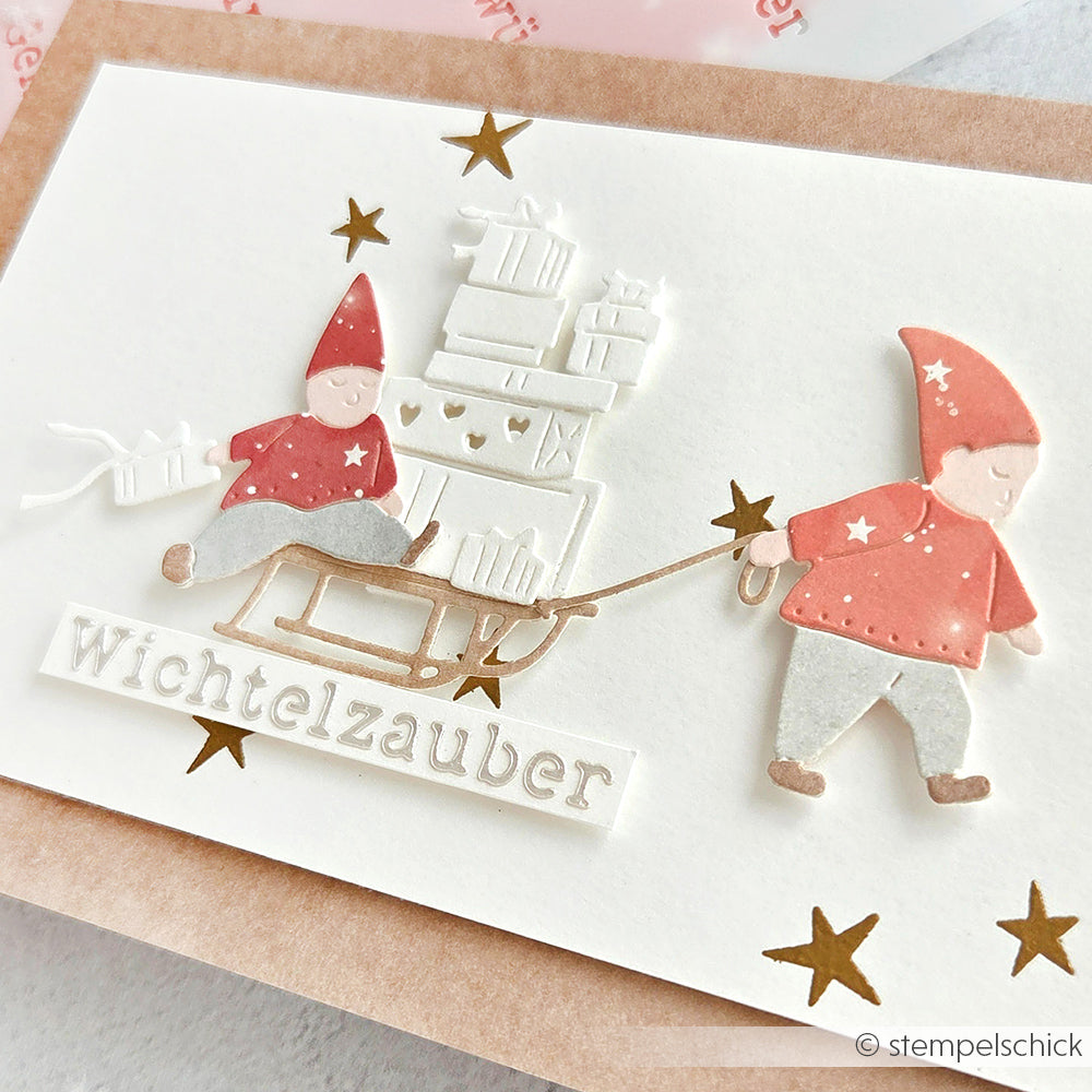 Zwei Wichtel mit Geschenken und einem Schlitten, goldenen Sternen und dem Text "Stencil Wichtelzauber" auf weißer Karte - ideal zum Basteln mit wiederverwendbaren Schablonen von Alexandra Renke.